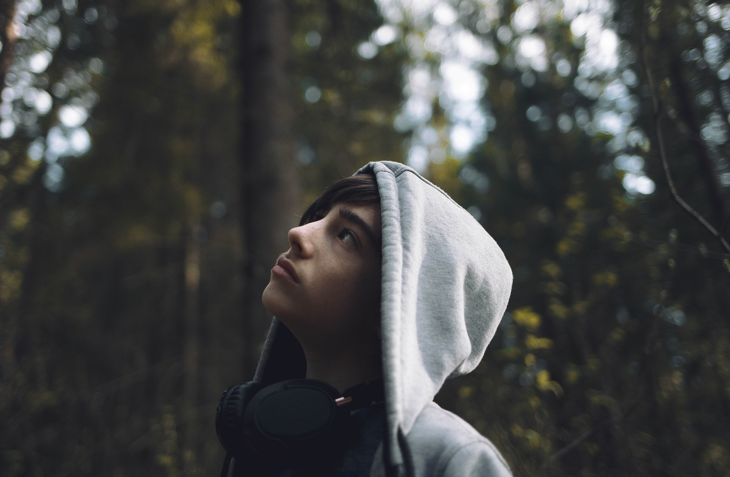 A young person in a grey hoodie gazes upward thoughtfully, with black headphones around their neck and trees blurred in the background.