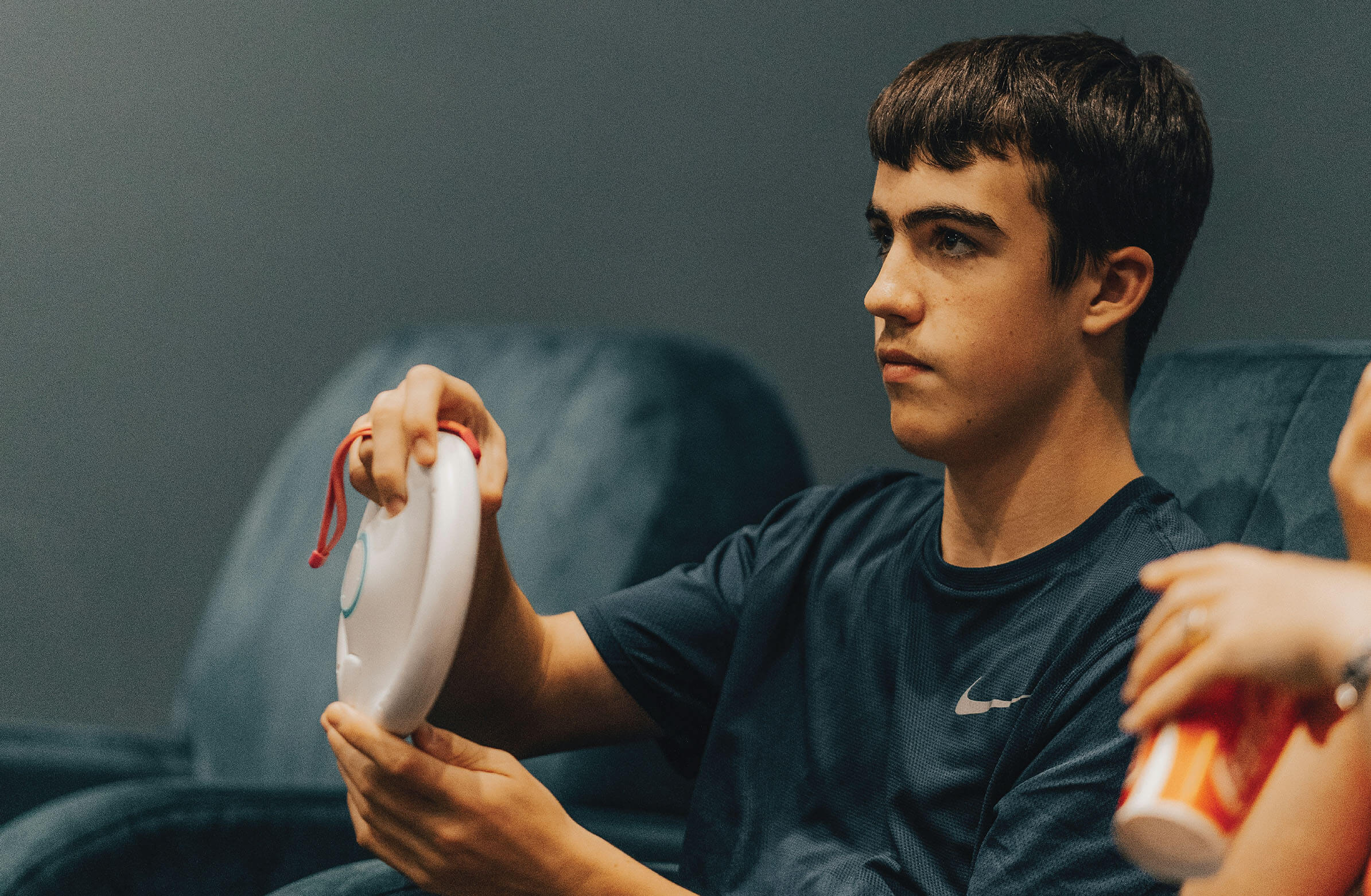 A teenage boy with short brown hair sits in a blue armchair, focused on holding a circular white object with a red strap in his hands. He is wearing a dark blue t-shirt