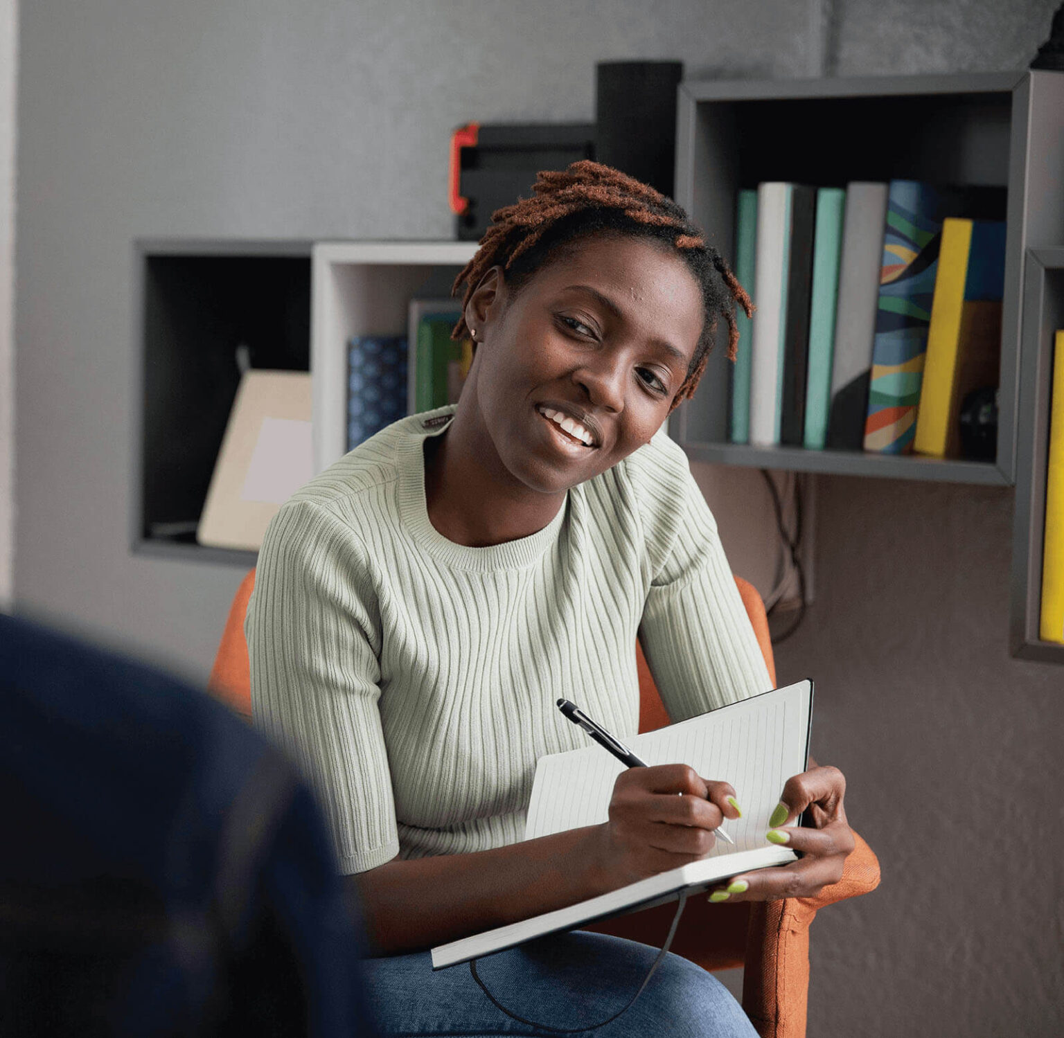 A smiling woman with short, curly hair, wearing a light green ribbed sweater, is seated indoors holding a notebook and pen. She appears engaged and is writing notes while looking towards someone out of the frame.
