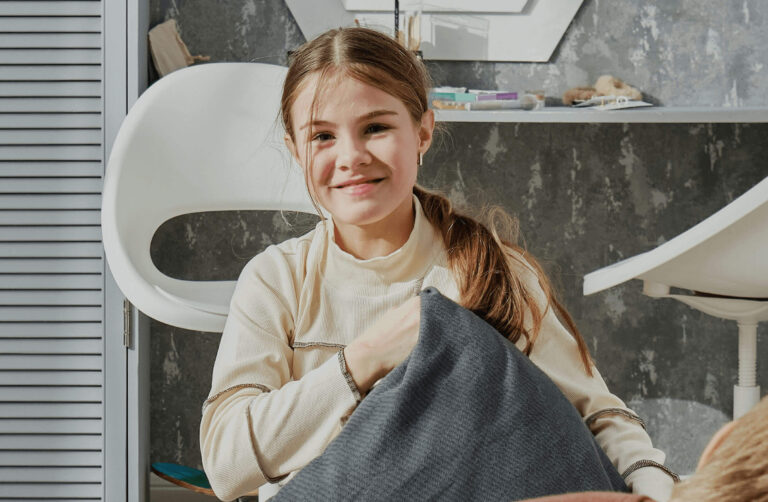 A young girl with long brown hair, tied in a ponytail, smiles while sitting on a chair in a modern room. She is wearing a light beige sweater and holding a cushion, with a white chair and shelves in the background.