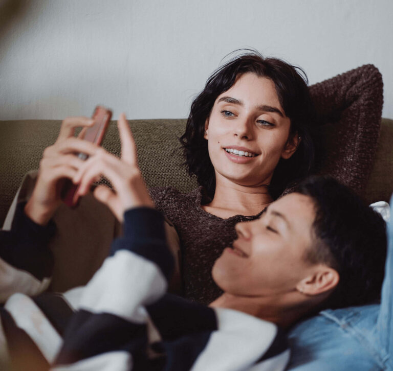 Two people are relaxing on a couch. The person on the left, with short black hair, is holding a smartphone and appears to be showing something on the screen to the other person, who is lying down with a contented expression. Both individuals are wearing casual clothing and seem to be enjoying a calm, comfortable moment together.
