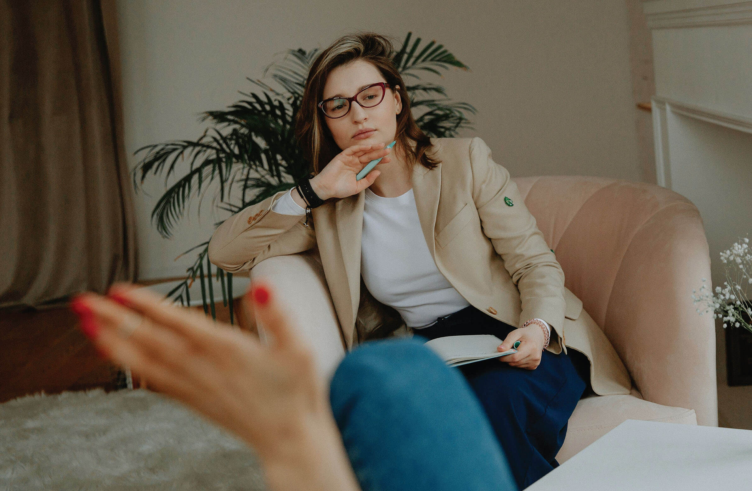 A woman wearing glasses and a beige blazer is sitting in a comfortable chair, attentively listening to someone off-camera. She has one hand on her chin and is holding a pen and notebook on her lap, suggesting she is a therapist or counselor. A plant is visible in the background, adding to the serene atmosphere of the room.
