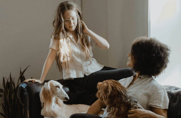 Two women are sitting together in a sunlit room. One woman with long, wavy hair and glasses is sitting on the arm of a black leather couch, while the other woman with curly hair is sitting on the couch, holding a small dog. A larger dog with long fur is lying next to them on the couch. Both women are smiling and appear relaxed.