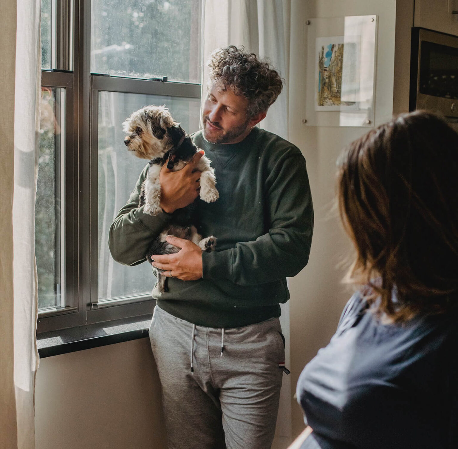 A man holding a small dog stands by a window, looking outside. Another person, partially visible from behind, faces the man, creating a warm, domestic scene filled with natural light.