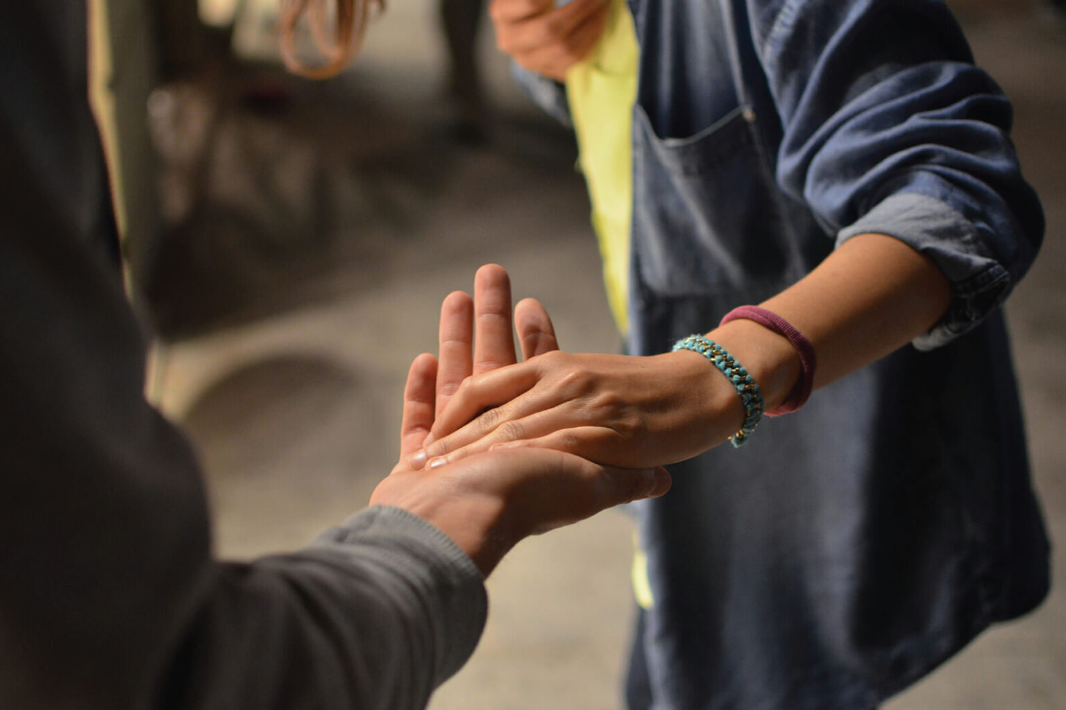 A close-up of two people holding hands. One person is wearing a denim shirt and has a bracelet on their wrist. The hands are gently touching in a supportive or comforting gesture.