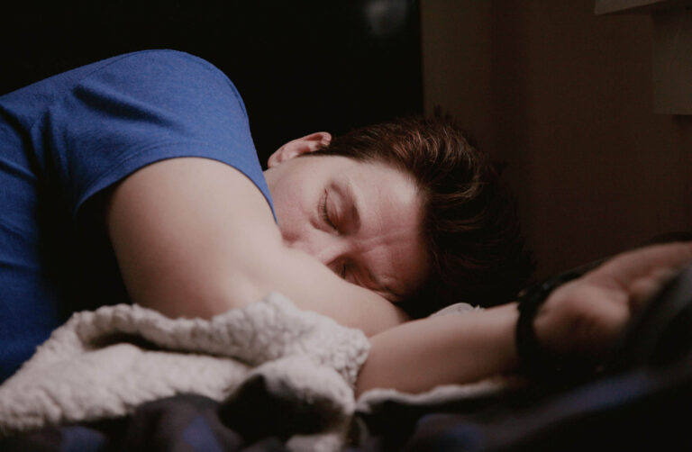 A person with short brown hair is lying on their side, asleep in a dimly lit room. They are wearing a blue shirt and resting their head on their arm, which is partially covered by a fluffy, light-colored blanket.