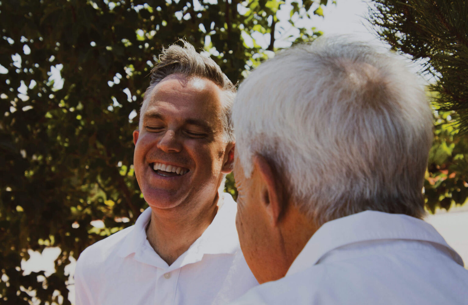 Two older men are outdoors, one with gray hair and the other with light brown hair. The man with light brown hair is smiling widely with his eyes closed, enjoying the conversation. The back of the head of the other man is visible as he faces the man who is smiling. The background includes green foliage in a sunlit setting.