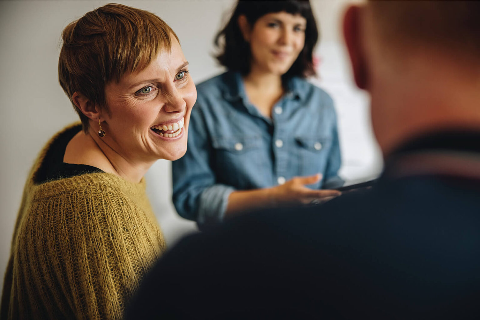 A woman with short light brown hair in a mustard sweater smiles while talking to two others. One, a dark-haired woman in a denim shirt, is blurred in the background, and the other is partially visible from behind