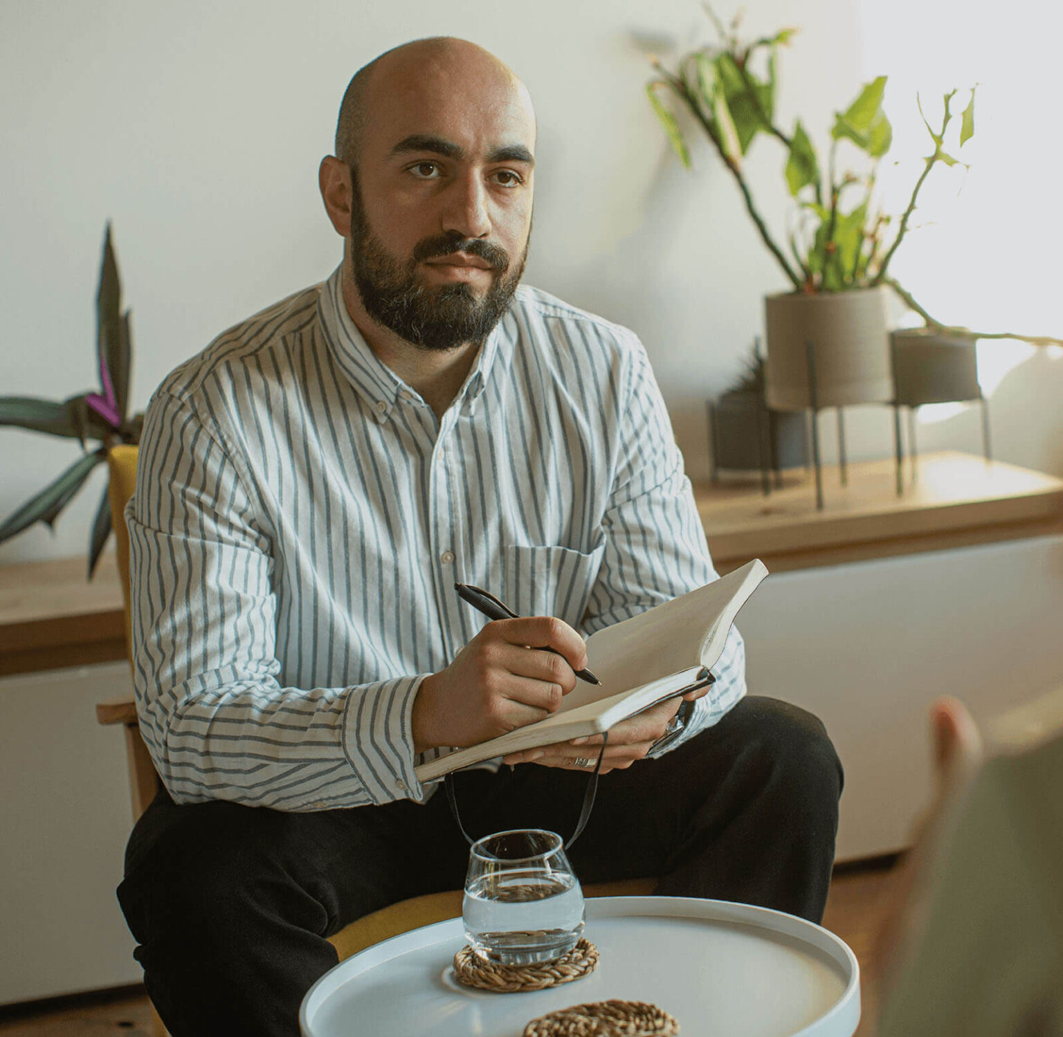 A bearded man with a bald head is seated indoors, holding a pen and writing in a notebook. He is wearing a white and blue striped shirt. A glass of water is placed on a small round table in front of him. The background includes some green plants, adding a natural touch to the scene.