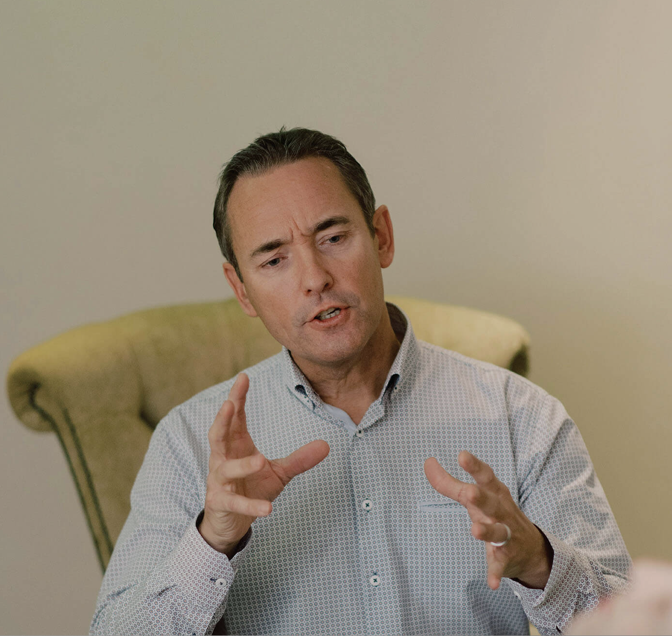A middle-aged man with short dark hair, wearing a light-colored, patterned shirt, is seated in a chair while speaking. His hands are gesturing expressively as he talks, indicating a thoughtful discussion. The background shows a light-colored wall and part of the upholstered chair.