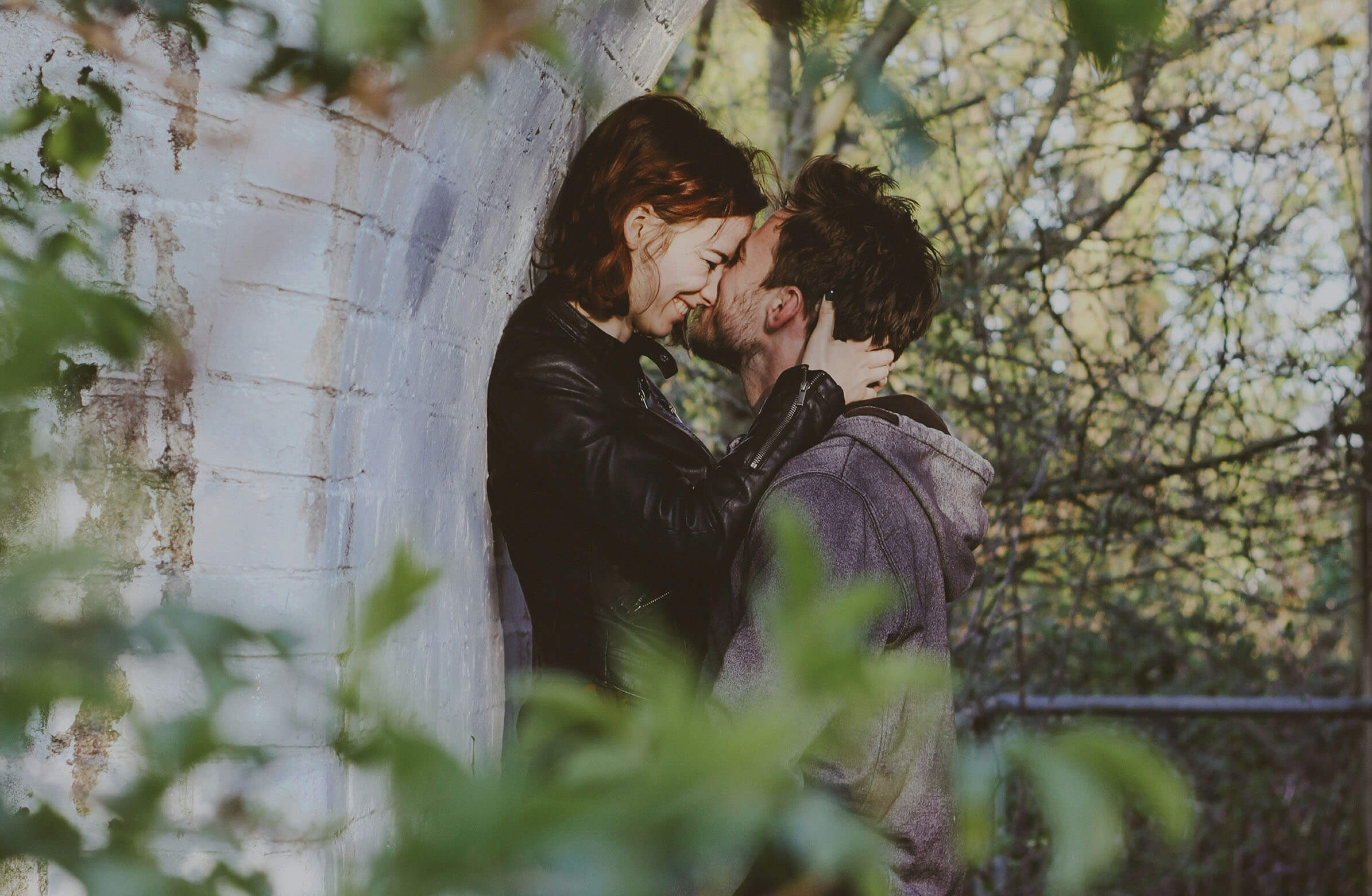 A couple sharing an intimate moment outdoors, with the woman leaning against a wall and the man close to her, both embracing affectionately amidst a backdrop of greenery.