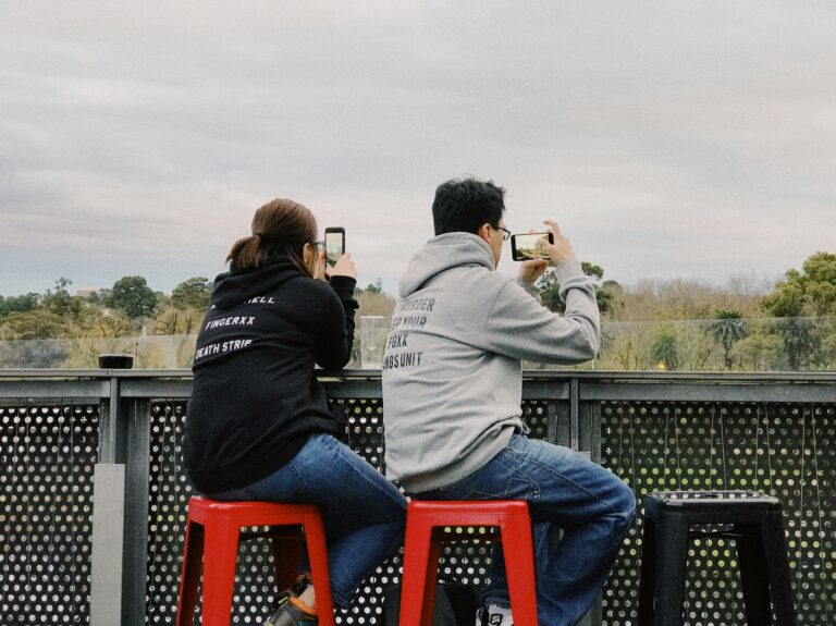 Two people are seated on red stools outdoors, facing away from the camera and taking photos with their smartphones. They are both wearing casual hoodies, with the person on the left in a black hoodie and the person on the right in a gray hoodie. The background features a scenic view with trees and a cloudy sky, suggesting that they are capturing a landscape or nature scene. The setting appears to be on a raised platform or balcony with a metal railing.
