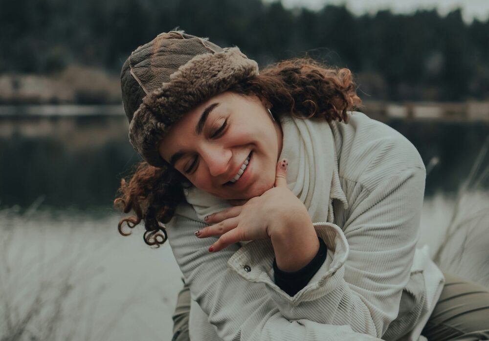 A young woman with curly hair smiles warmly, eyes closed, wearing a fur-lined hat and scarf by a calm outdoor setting with water and trees.