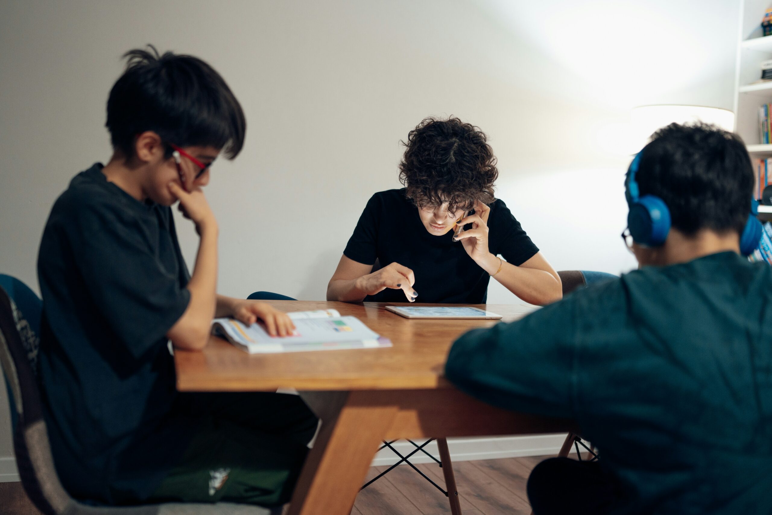 Three people sit around a wooden table in a well-lit room: one reads a book, another uses a tablet while on the phone, and the third, wearing blue headphones, looks at the table.