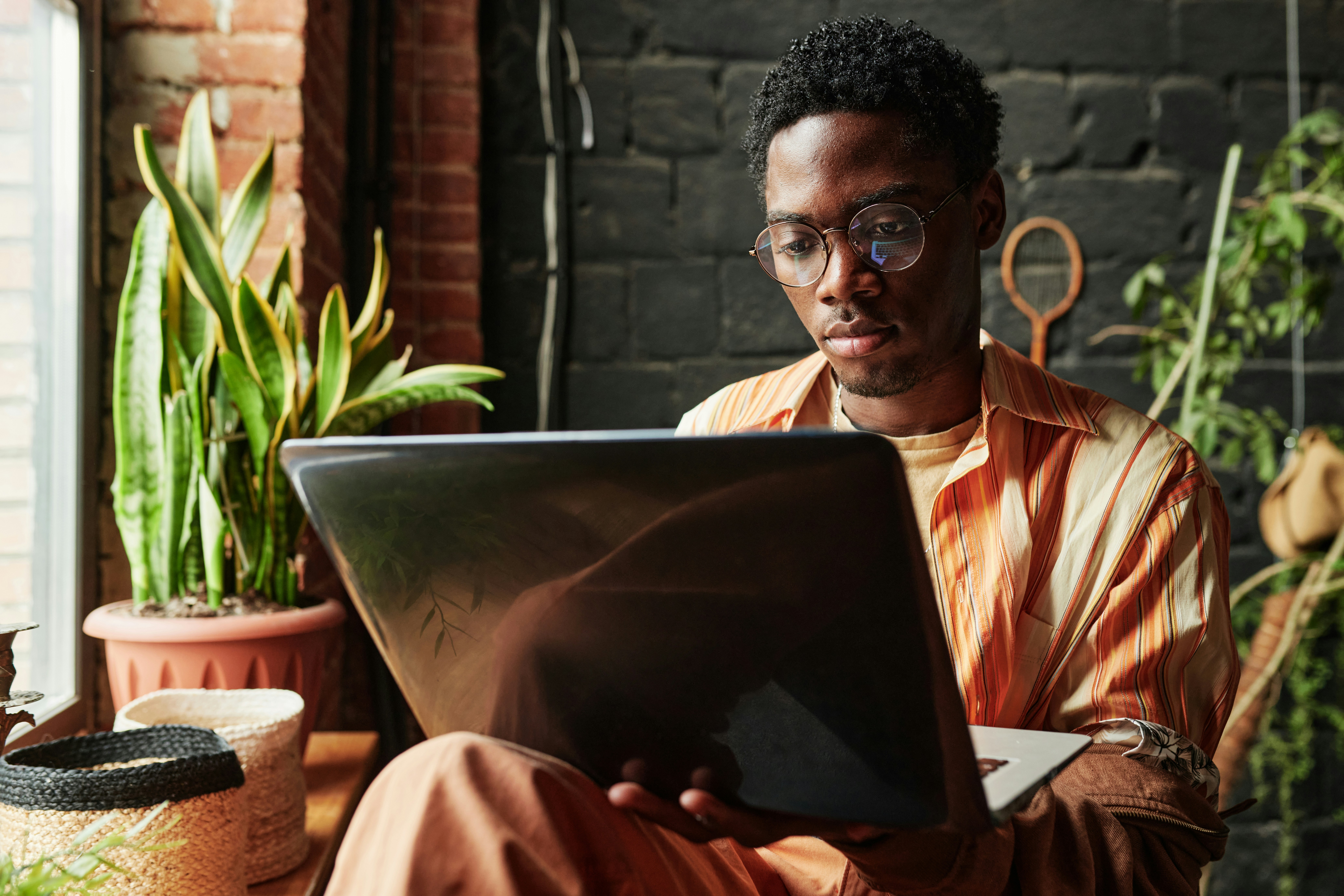 A man wearing glasses and a striped shirt is focused on working on a laptop, surrounded by indoor plants and natural decor.