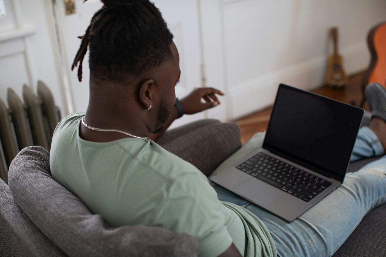 A man with short dreadlocks, wearing a light green t-shirt, relaxing on a sofa while using a laptop with a blank screen, in a cosy room with a guitar in the background.