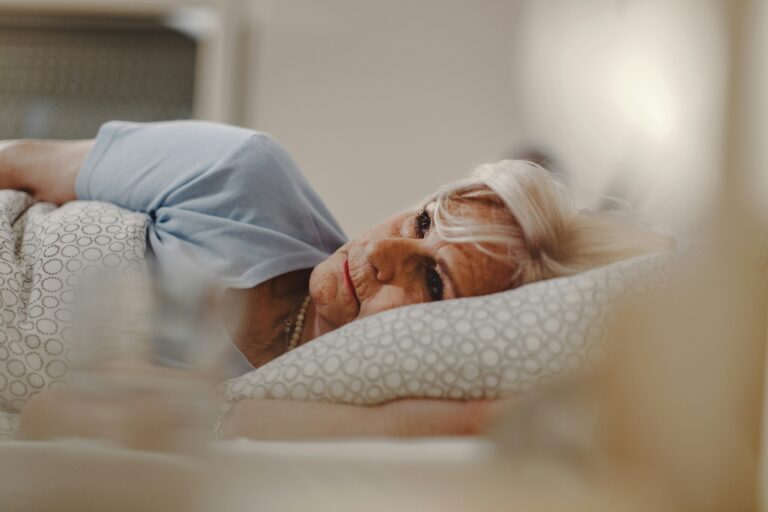 Elderly woman with light hair lying on a pillow, looking thoughtful with a gentle expression